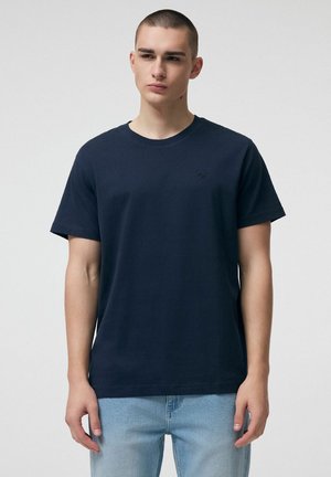 Młody mężczyzna z krótkimi włosami, ubrany w gładką ciemnogranatową koszulkę i jasnoniebieskie dżinsy, stoi na jednolitym tle.