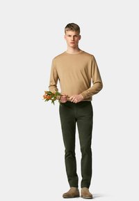 Lichtbruin langemouw overhemd gecombineerd met donkergroene aansluitende broek. Het model houdt een tak met oranje bloemen vast en draagt tan enkellaarsjes.