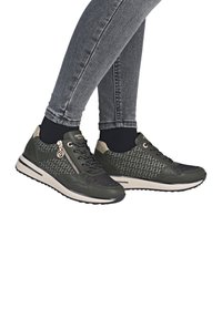 Mørkegrønne sneakers med mønster, snørebånd og lynlås på siden, båret med sorte sokker og grå tætsiddende jeans mod hvid baggrund.