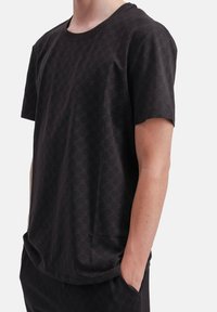 Man in een zwart gemusterd t-shirt met korte mouwen en bijpassende broek, met de rechterhand in de broekzak, tegen een witte achtergrond.