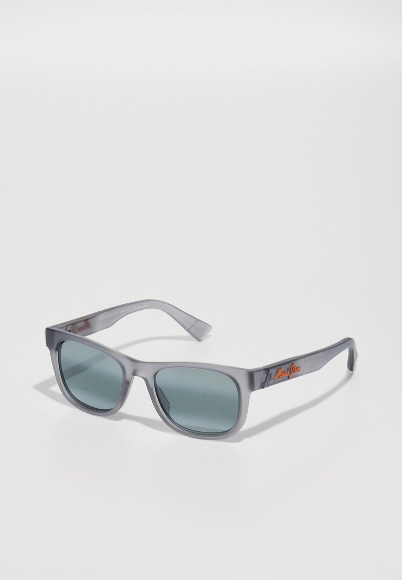 Gafas de sol de plástico gris con lentes tintados grises. Montura cuadrada con acabado mate. Presenta un logo naranja en la patilla.