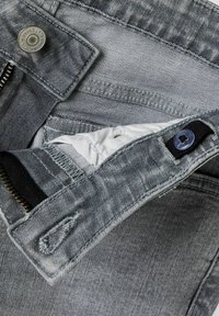Jeans en denim gris avec une taille déboutonnée montrant une bande élastique réglable avec un bouton et une fermeture éclair visible en dessous.