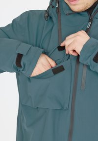 Tealinvärinen vedenpitävä takki, jossa on säädettävä huppu. Varustettu etutaskulla, jossa on tarrakiinnitys ja vetoketjulliset ilmanvaihtoaukot. Sujuva pinta ja rento malli.