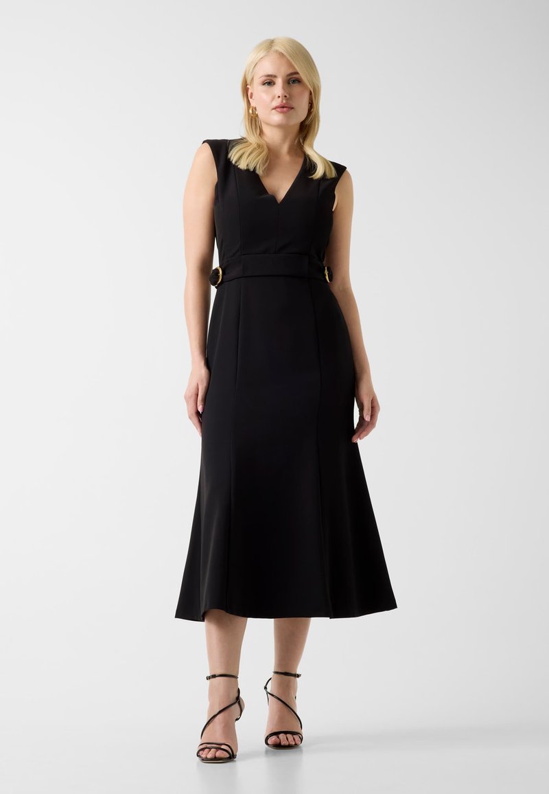 Femme blonde portant une robe midi noire sans manches avec un décolleté en V et une taille ceinturée, associée à des sandales noires à talons hauts et à brides.