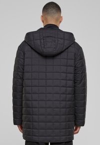 Svart kviltet pufferjakke med hette, med en boksaktig design og teksturert overflate. Ingen synlig maskinvare. Mid-lengde snitt.