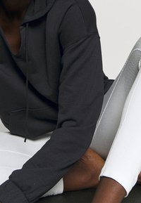 Felpa nera con cappuccio e vestibilità ampia, dotata di una tasca frontale e sottili dettagli di cucitura. Indossata con leggings bianchi, che evidenziano il contrasto.