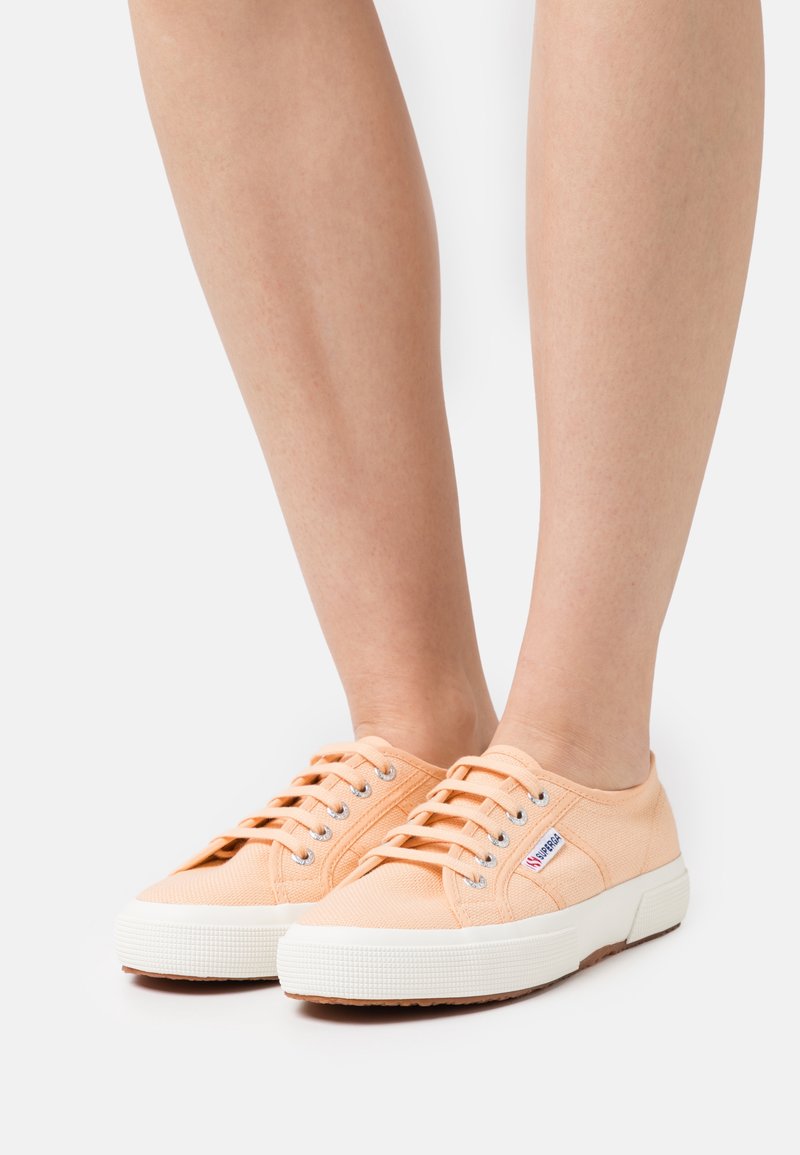 Paire de baskets en toile couleur pêche avec semelles en caoutchouc blanches et lacets, portées pieds nus, posées sur une surface blanche.