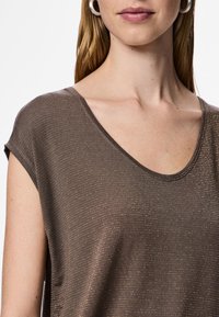 Ruskea, lyhythihainen paita, jossa on V-kaula-aukko. Teksturoitu kangas, jossa hienovaraiset kultaiset raidat. Rentoutunut malli korostaa kaula-aukkoa ja hartioita.