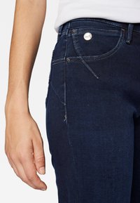 Vaqueros de mezclilla azul oscuro con un diseño ajustado, detalles de costura visibles y acentos de botones metálicos. Bolsillo lateral y un ligero deshacimiento en el dobladillo.