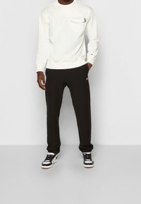 Felpa bianca con taschino sul petto e pantaloni della tuta neri con logo. Scarpe da ginnastica nere e bianche completano il look.