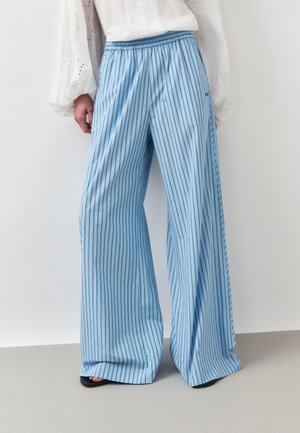 Person trägt hellblaue, weit geschnittene Hosen mit vertikalen dunklen Streifen, kombiniert mit einer weißen Bluse mit langen Ärmeln und schwarzen offenen Schuhen.