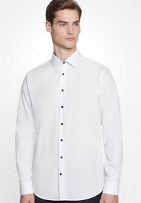 Weißes Langarmhemd mit Button-Down-Kragen, das schwarze Knöpfe entlang der vorderen Knopfleiste aufweist und eine glatte Stofftextur hat.