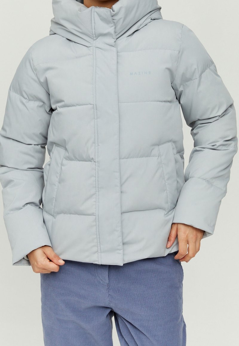 Personne portant une doudoune gris clair avec capuche et un pantalon en velours bleu, mains près des poches de la veste.