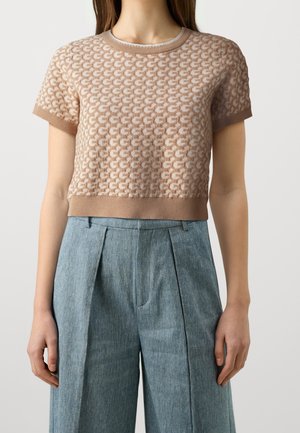 Frau trägt ein beiges, kurzärmeliges, gemustertes Stricktop kombiniert mit hoch taillierten, weiten blauen Hosen vor einfachem Hintergrund.
