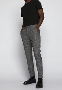 Šedé kárované kalhoty s úpravou na míru, vyrobené z texturované látky. Kombinovány s černou košilí s krátkým rukávem a černými botami.