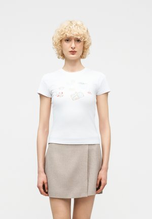 Ξανθός/ή φοράει λευκό μπλουζάκι με γραφικό σχέδιο και μπεζ μίνι φούστα, στέκεται μπροστά σε απλό φωτεινό φόντο, κοιτάζοντας προς τα εμπρός.