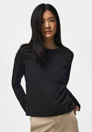 Jeune femme aux longs cheveux foncés portant une chemise noire à manches longues et un pantalon beige, debout devant un fond clair uni.