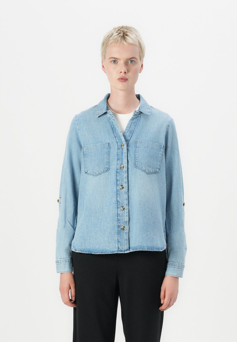 Camisa de ganga azul clara com mangas longas, fecho de botões, dois bolsos no peito e mangas dobráveis. Textura suave e design de colarinho clássico.
