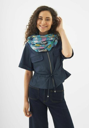 Femme souriante aux cheveux bouclés portant un haut zippé bleu foncé et un pantalon, accessoirisée d'une écharpe colorée imprimée de dessins animés autour du cou.