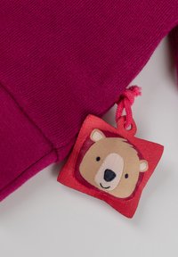 Pullover fucsia con finitura testurizzata, caratterizzato da un'etichetta cucita che raffigura il volto di un orso in colori tenui, attaccata con un anello rosa.