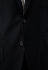 Nærbillede af en mørkeblå blazer med to sorte knapper knappet, der viser forlommerne og en del af en hvid skjorte under.