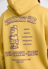Gelber Kapuzenpullover mit lila grafischem Design, das eine Zeichentrickfigur und Text zeigt, der Artikel wie Getränke, Snacks und Zeitschriften auflistet.