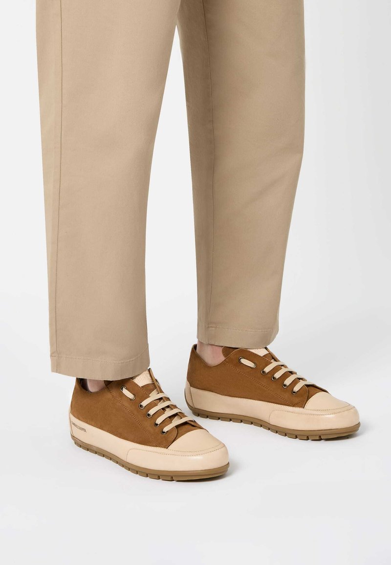 Sneakers in camoscio marrone con suole e punte in gomma beige, dotati di lacci beige e sottili perforazioni, abbinati a pantaloni beige chiaro.