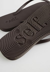 Brązowe klapki z fakturowanymi wkładkami i podeszwami z wzorem fal, z wypukłym napisem „self.” na wkładkach i podeszwach.