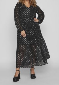 Vestido negro transparente con patrones en forma de hoja blanca, mangas largas, escote en V y dobladillo en capas. Combinado con zapatos de plataforma negros.