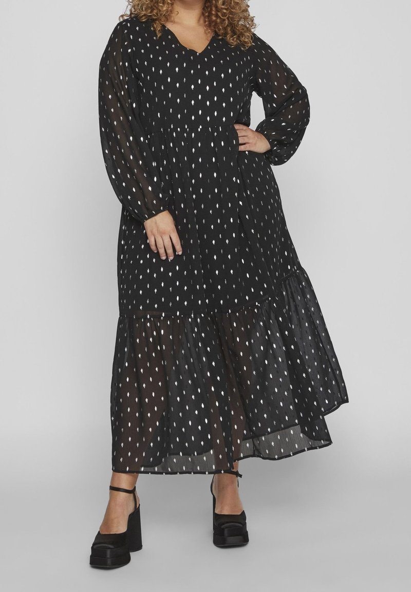 Vestido negro transparente con patrones en forma de hoja blanca, mangas largas, escote en V y dobladillo en capas. Combinado con zapatos de plataforma negros.