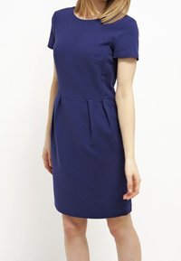 Kvinna som bär en figurnära, kortärmad, knälång klänning i marinblått med subtila veck vid midjan mot en enfärgad bakgrund.