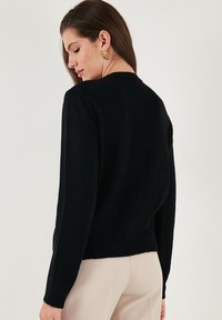 Svart stickad tröja med ribbad fåll och muddar, som har rund halsringning och långa ärmar, sedd bakifrån.