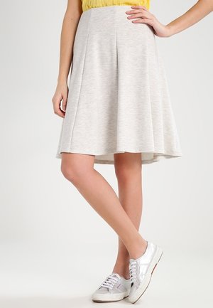 Femme portant une jupe trapèze gris clair plissée, associée à des baskets argentées et un haut jaune, debout avec une main sur la hanche.