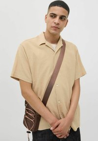 Camisa corta de manga corta en tono beige con una tela texturizada, diseño con cuello y dos botones en la parte delantera, combinada con un bolso cruzado marrón.