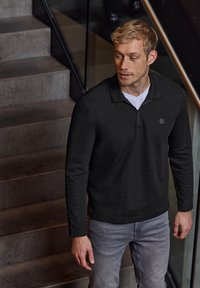 Svart zip-up tröja med lång ärm, med krage och en liten logotyp. Bärs över en vit t-shirt, ihop med ljusgrå jeans.