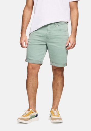 Hombre con pantalones cortos enrollados de color verde claro, camiseta blanca y zapatillas blancas con detalles en amarillo, verde y azul marino, de pie sobre un fondo liso.