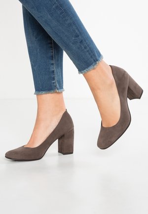 Escarpins à talons hauts en daim marron avec un bout rond et un talon épais, associés à un jean à ourlet effiloché. Texture lisse, design minimaliste.