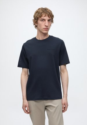 Jeune homme aux cheveux blonds bouclés portant un t-shirt bleu marine uni et un pantalon beige, debout devant un fond blanc.