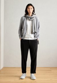 Cardigan gri tricotat peste un tricou alb, asortat cu pantaloni negri și adidași gri, stând pe un podea din lemn pe un fundal simplu.