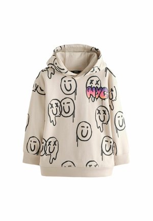 Beige hættetrøje med sorte smiley-tegninger, nogle smeltende eller overstregede, og farverig "NYC" tekst på brystet, båret udendørs nær et grønt hegn.
