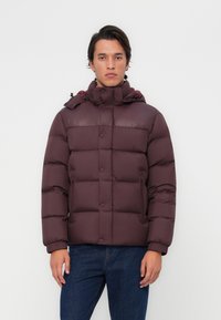 Bordo puffer jakna s visokim ovratnikom, patentnim zatvaračem i gumbom. Sadrži quilted dizajn i kapuljaču s podesivim vezicama.