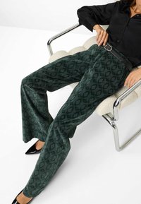 Pantaloni a gamba larga con motivo verde, realizzati in tessuto morbido, caratterizzati da una texture sottile e abbinati a una cintura nera e a una camicia nera.