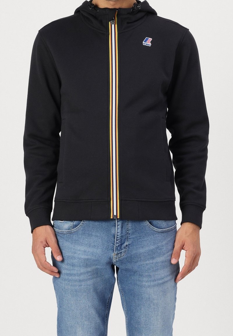 Uomo che indossa una felpa nera con zip, dotata di una cerniera a righe multicolori e un piccolo logo sul petto, abbinata a jeans azzurri.