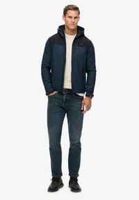 Dunkelblaue Jacke mit Reißverschluss und Kapuze, kombiniert mit schwarzen Schultern, vorderer Reißverschlusstasche, getragen mit leichtem Pullover, blauen Jeans und schwarzen Schuhen.