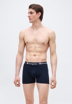 Άνδρας χωρίς πουκάμισο με κοντά καστανά μαλλιά, φορώντας μαύρα εσώρουχα Champion boxer briefs, στέκεται μπροστά σε λευκό φόντο, κοιτώντας προς τα αριστερά.