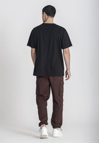 Černé bavlněné tričko v kombinaci s hnědými cargo kalhotami s bočními kapsami, doplněné o bílé tenisky. Model je viděn zezadu.