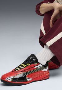 Scarpe sportive rosse e nere con lacci gialli, dettagli bianchi e fori di ventilazione. Indossate con calze bianche e pantaloni bordeaux con strisce bianche.