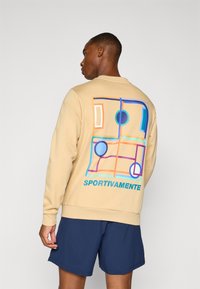 Beige sweatshirt med fargerik grafisk design på baksiden, med abstrakte former og teksten "SPORTIVAMENTE" i blått. Myk tekstur.