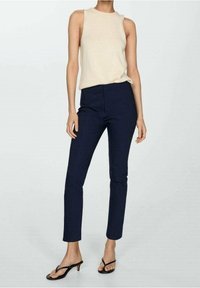 Donna che indossa un top senza maniche beige, pantaloni blu scuro aderenti e sandali neri con tacco, in piedi contro uno sfondo chiaro uniforme.