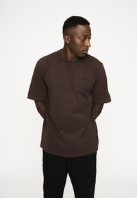 Brun bomulds t-shirt med rund halsudskæring og korte ærmer. Har et diskret logo på brystet. Bæres med mørke bukser, generelt simpelt design.
